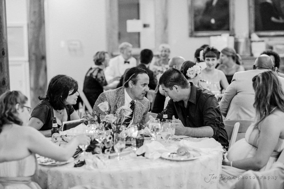 A black and white photo of a formal event. People sit around a table with drinks and food, engaged in conversation. The room is lively, with other tables and guests in the background. Theres a sense of camaraderie and celebration.