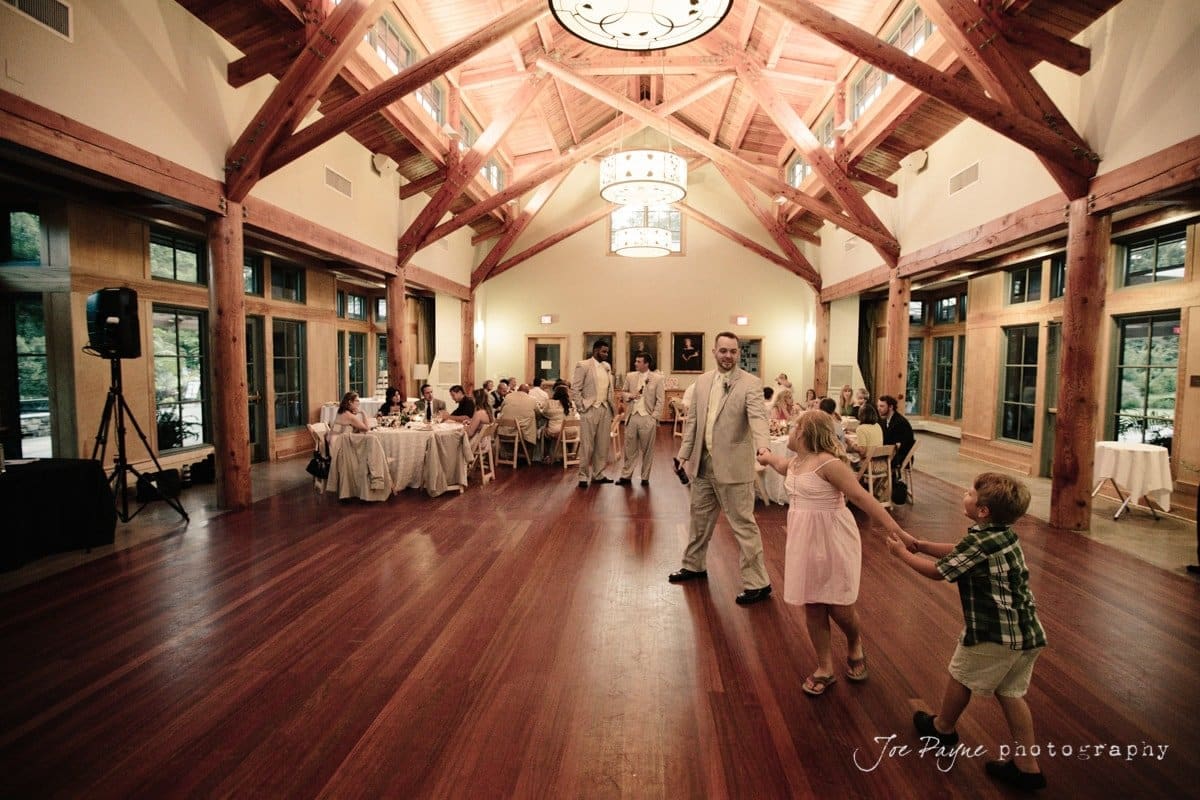 A spacious wooden hall with a high ceiling is set for a formal event. Guests are seated at round tables. In the foreground, a man in a suit is playfully dancing with two children. Natural light streams through large windows on the sides.