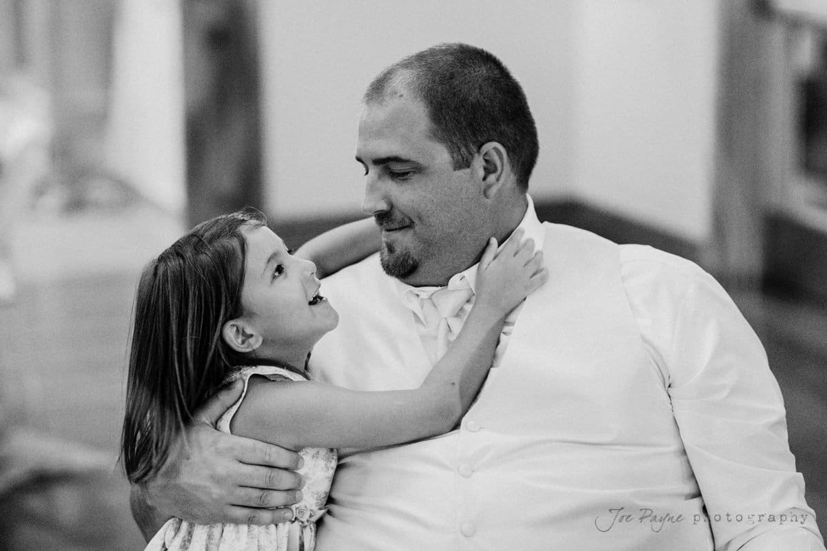 A black and white photo of a smiling man sitting while a young girl hugs him. The girl looks up affectionately at the man, who is wearing a vest and tie. The background is softly blurred.