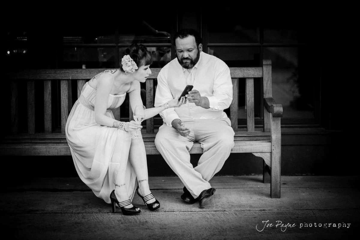 A woman in a dress and a man in a shirt and pants sit on a wooden bench, looking at a phone together. The womans flower hair accessory and the black-and-white color scheme add a timeless feel to the scene.