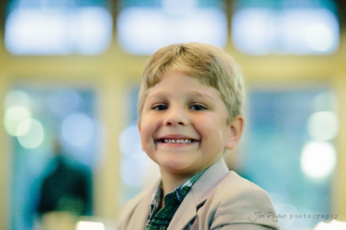 A young boy with short blond hair smiles brightly while wearing a light-colored blazer over a plaid shirt. The background is softly blurred, suggesting an indoor setting with natural light.