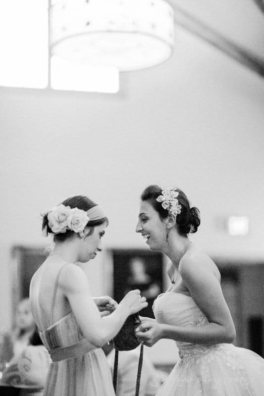 Two women in formal dresses smiling and interacting closely in a warmly lit room. Both have hair adorned with flowers; one is in a strapless gown. The photo captures a candid, joyful moment.