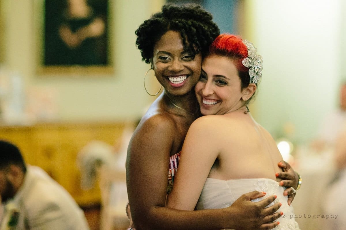 Two women smiling and hugging at an indoor gathering. One wears a colorful dress, and the other is in a strapless white dress with a sparkly hair accessory. The background is softly blurred, with a painting on the wall.