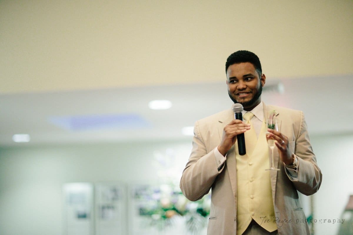 A man in a beige suit and yellow vest holds a microphone and a champagne flute, smiling. He stands in a softly lit room with blurred background elements.