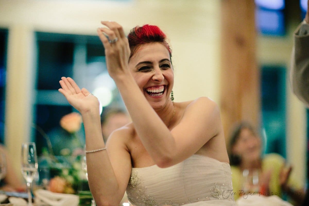 A woman with short red hair is sitting at a table, smiling and raising her hands in a lively gesture. She is wearing a strapless dress, and there are blurred people and flowers in the background, suggesting a celebratory event.