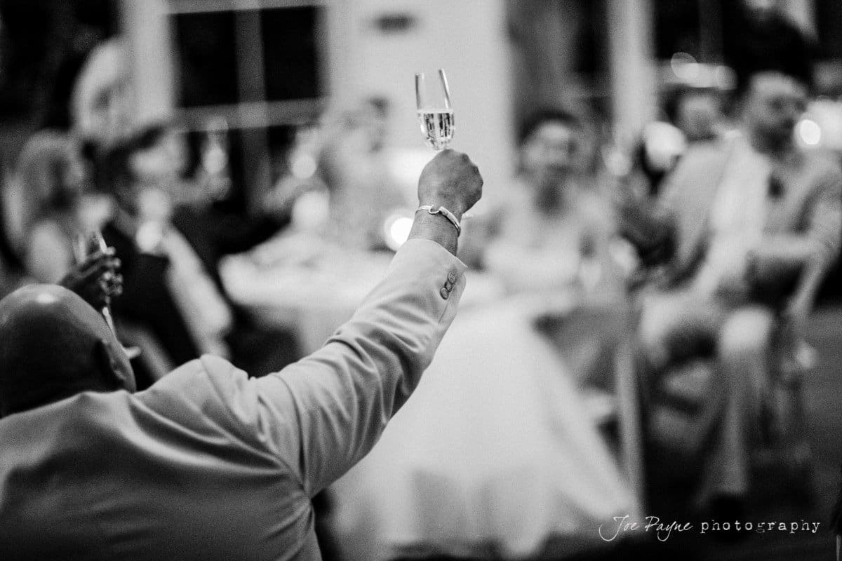 A person in a suit raises a glass in a toast at a formal gathering. Blurred guests sit around a table in the background. The scene is in black and white, with a sense of celebration and camaraderie.
