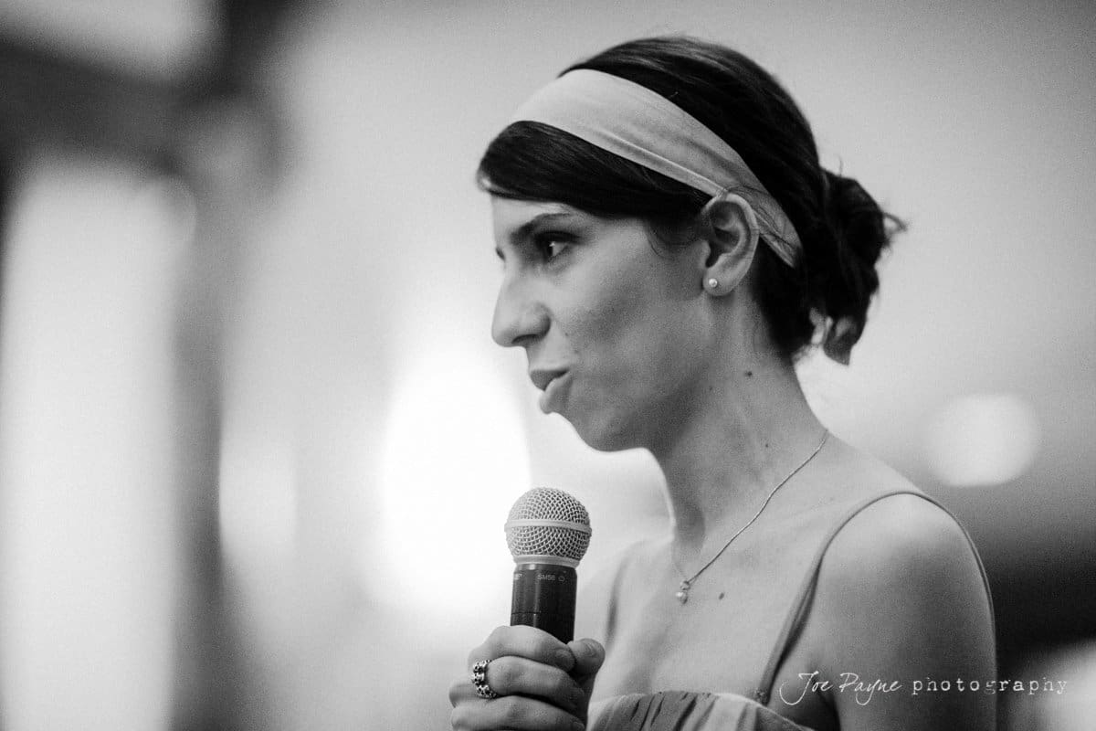 A woman with a headband, holding a microphone, is speaking. The setting appears to be indoors, with blurred lights in the background. The image is in black and white.