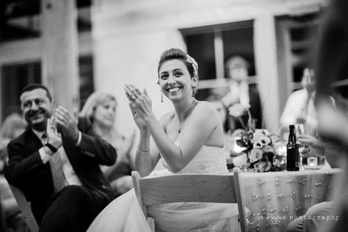 A black and white photo of a bride in a strapless dress sitting and clapping at a wedding reception. Shes smiling and looking to the side. Other guests, also clapping, are visible in the background with floral decorations on the table.