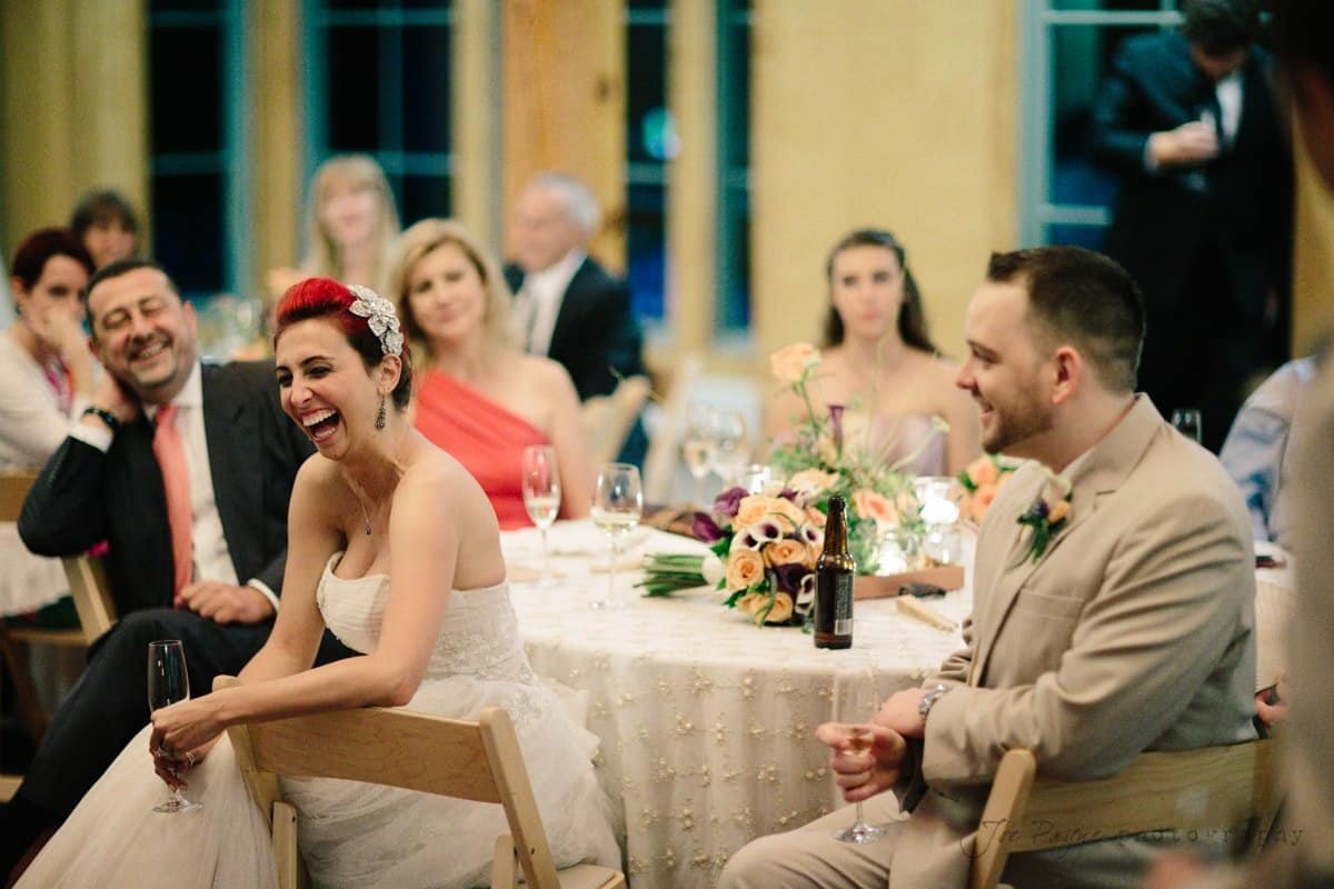 A bride with red hair in a white dress laughs joyfully, surrounded by seated guests, including a man in a beige suit. The table is decorated with flowers and glasses, creating a warm, celebratory atmosphere indoors.
