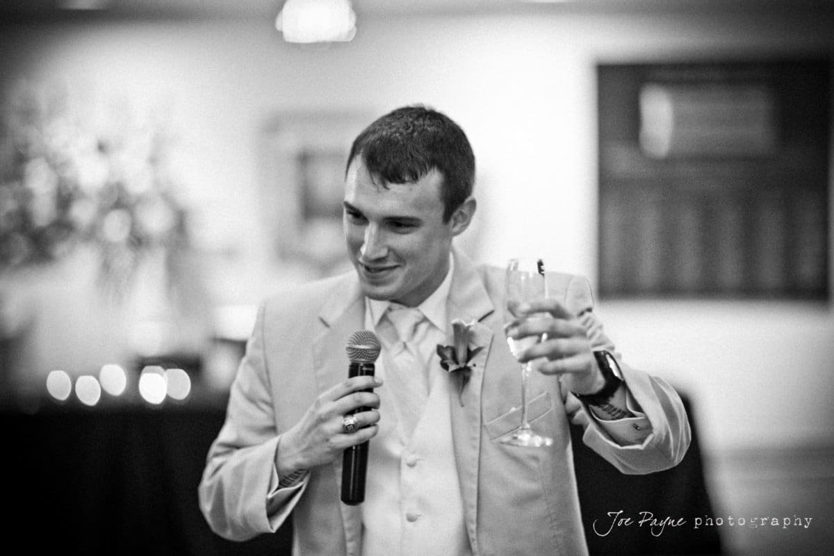 A man in a suit holds a microphone and raises a wine glass, as if giving a toast. The background is softly blurred, suggesting a formal event or celebration.