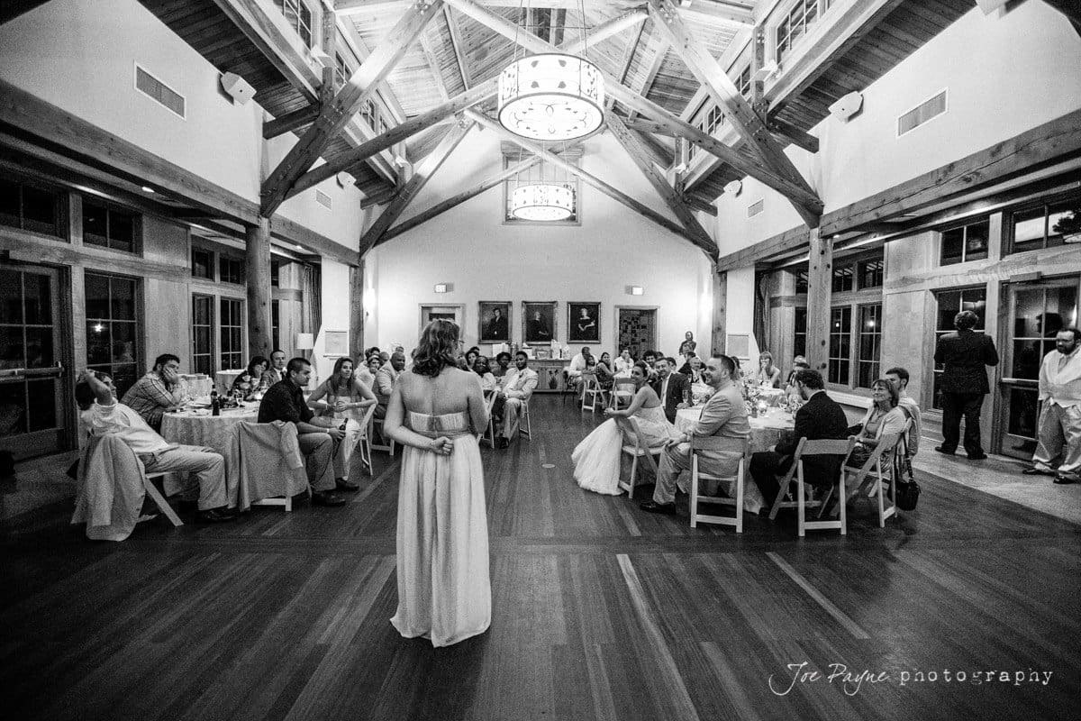 A black and white image of a wedding reception in a large hall with wooden beams. Guests are seated at round tables, and a woman stands in the center, facing them. A chandelier hangs from the high ceiling.