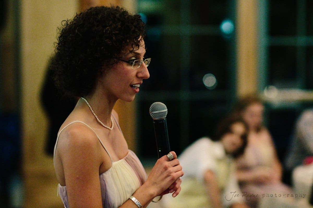 A woman in a sleeveless dress and glasses is speaking into a microphone. She has curly hair and is wearing a necklace and ring. The background is softly blurred, with people seated and windows visible.