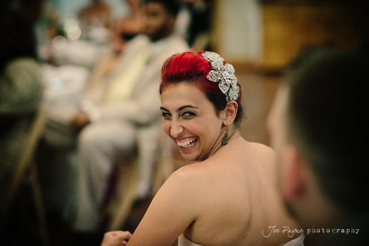 A woman with bright red hair adorned with an ornate hairpiece is looking back and smiling broadly. She is wearing a white dress, and people are seated in the background, slightly blurred.