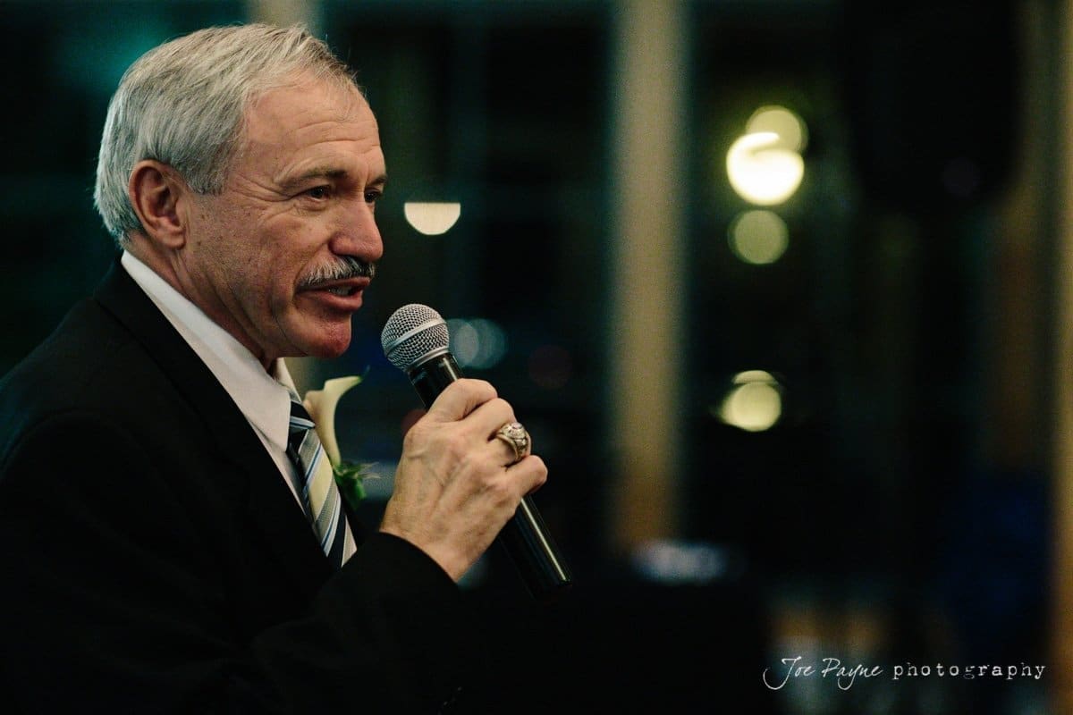 An older man with a mustache is speaking into a microphone. He is wearing a dark suit and a ring on his right hand. The background is blurred with soft lighting.
