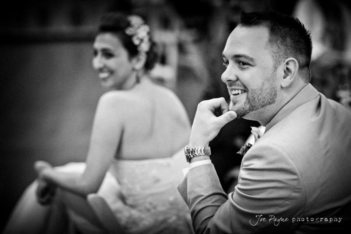 A black and white photo of a smiling man in a suit with a watch, sitting with his hand on his chin. In the background, a woman in a wedding dress appears joyful, slightly out of focus.