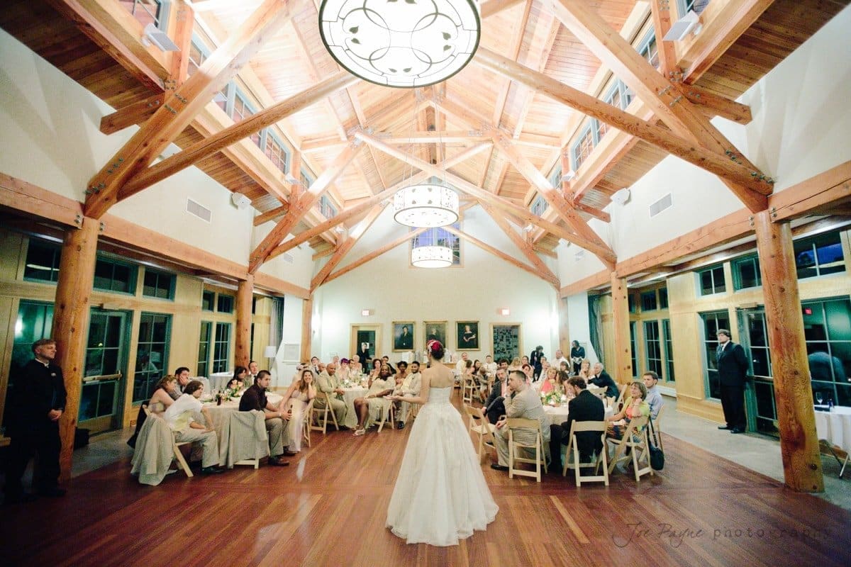 A bride in a white gown stands in a spacious, wood-beamed hall with a high ceiling, addressing guests seated at round tables. A large, circular light fixture hangs from the ceiling, and natural light streams through tall windows.