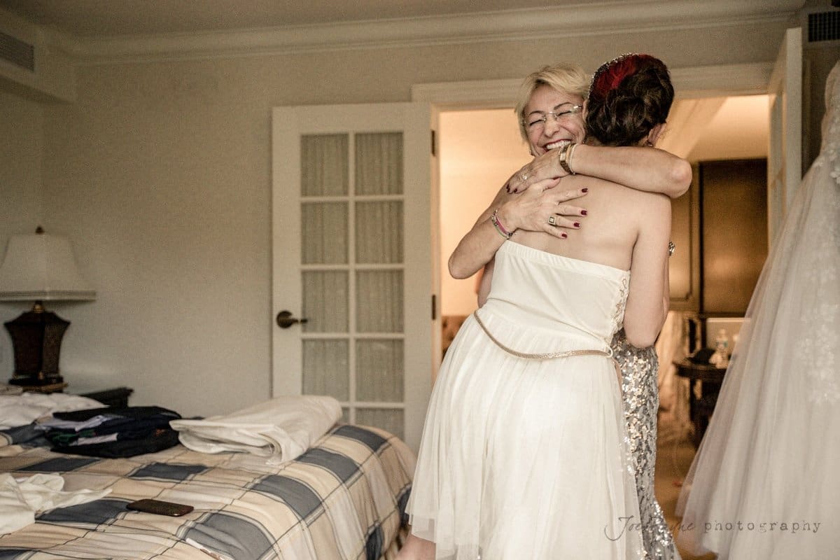 Two women share a warm hug in a softly lit bedroom. One wears a strapless dress, while the other wears a sparkling gown. A wedding dress hangs on the right, and a bed with folded clothes is on the left.