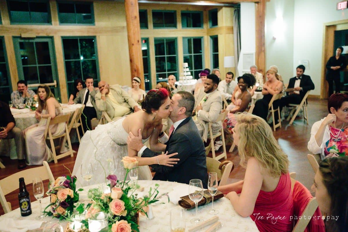 A bride leans over to kiss a seated man during a lively wedding reception. Guests are seated around tables decorated with flowers, plates, and glasses. Some people are watching the couple, while others are engaged in conversation.