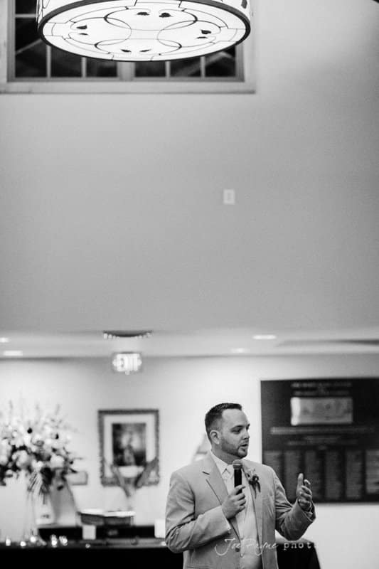 A man in a suit holds a microphone, speaking in a room with a high ceiling. A large, circular light fixture hangs above. In the background, there are flowers on a table and framed items on the wall. The setting appears to be formal.