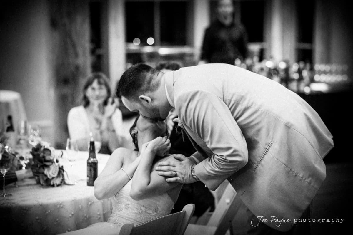 A groom in a suit affectionately kisses a seated bride, who is wearing a wedding dress, on the forehead. Guests at nearby tables watch the couple, smiling. The setting appears to be a warmly lit reception hall.