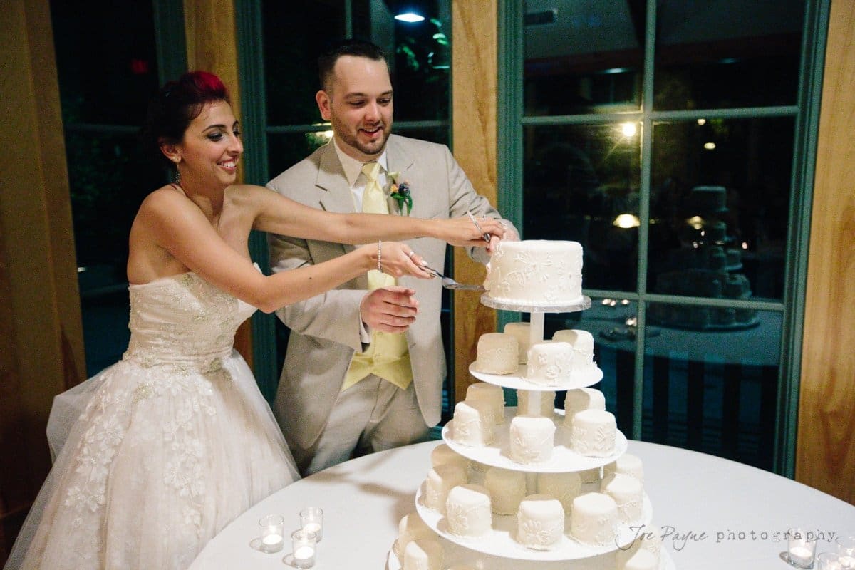 A bride in a white gown and a groom in a light-colored suit cut an elegant tiered white cake together. The cake is decorated with intricate designs, and small candles are lit on the table nearby. The room is softly lit.