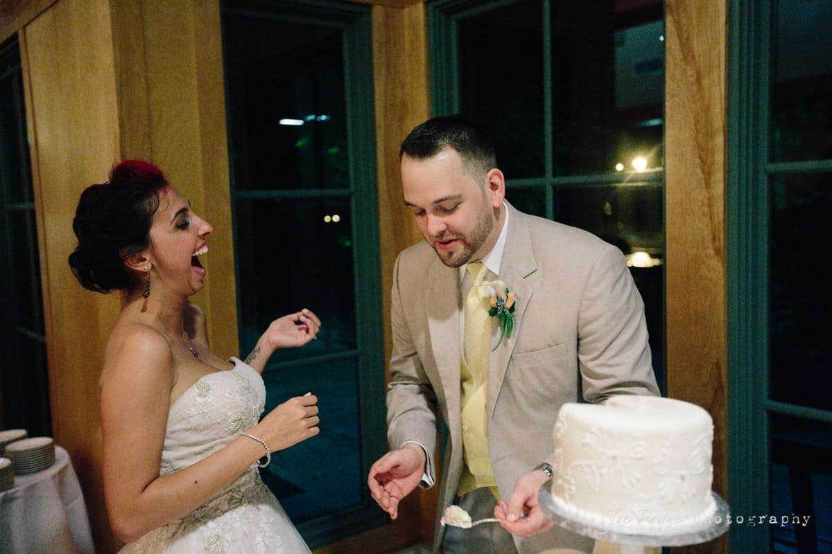 A bride and groom laugh together as they stand in front of a decorated cake. The bride is wearing a strapless gown and the groom is in a beige suit with a yellow tie. They appear to be sharing a joyful moment indoors.