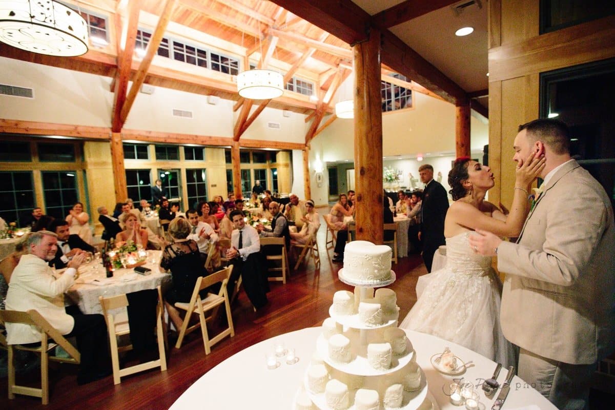 A bride and groom playfully feed each other cake in a large, warmly lit reception hall. Guests seated at tables watch and laugh. A tiered white wedding cake is visible in the foreground.