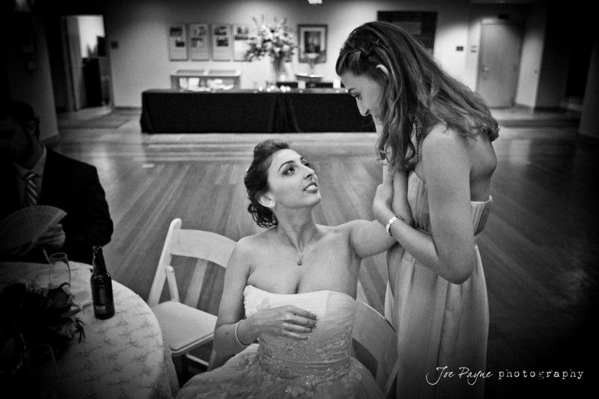 A black-and-white image of two women. One in a wedding dress is seated, and the other is standing, holding the seated womans hand. They are engaged in conversation in a room adorned with floral arrangements.