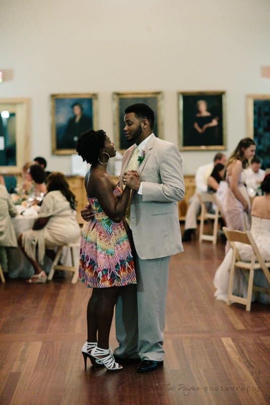 A couple dances together at a formal event. The man is in a light suit and the woman is in a colorful dress, both looking into each others eyes. People are seated in the background, and framed portraits are visible on the walls.