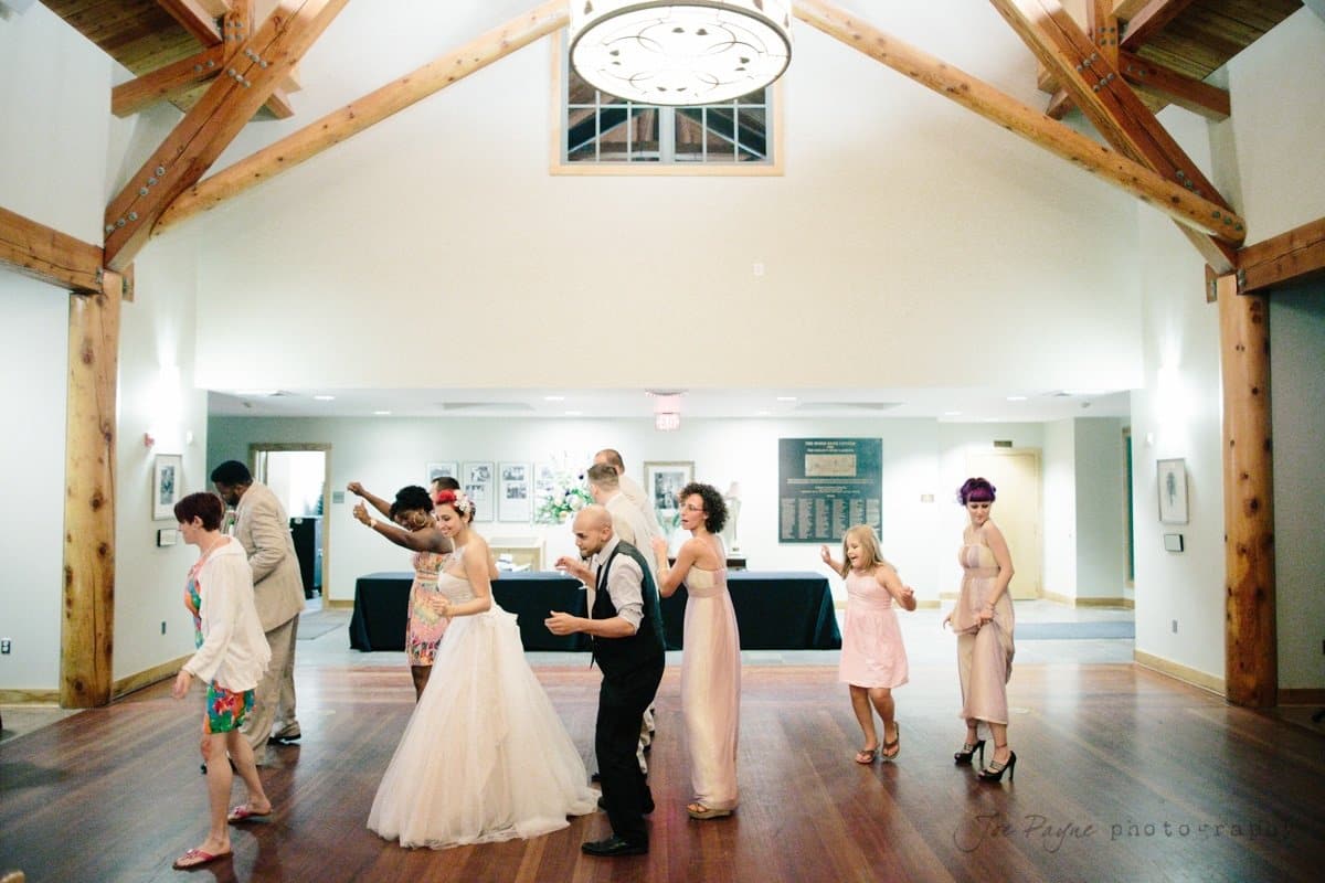 A group of people, including a bride in a white dress, are dancing in a brightly lit room with wooden beams. The setting appears to be a wedding reception, with guests dressed in formal and colorful attire.