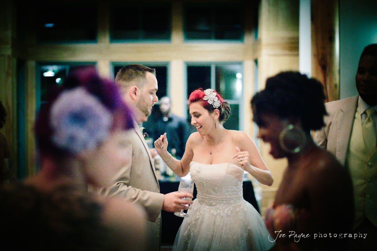 A bride in a strapless white gown dances energetically with guests at a wedding reception. She has flowers in her hair and holds a drink. People around her are smiling and dressed in formal attire. The room has large windows and warm lighting.