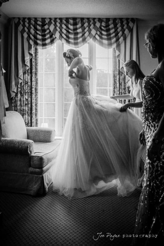 A black and white photo shows a bride in a wedding gown standing near a window as two people assist her with the dress. The scene is set in a room with a patterned curtain and a plush chair.