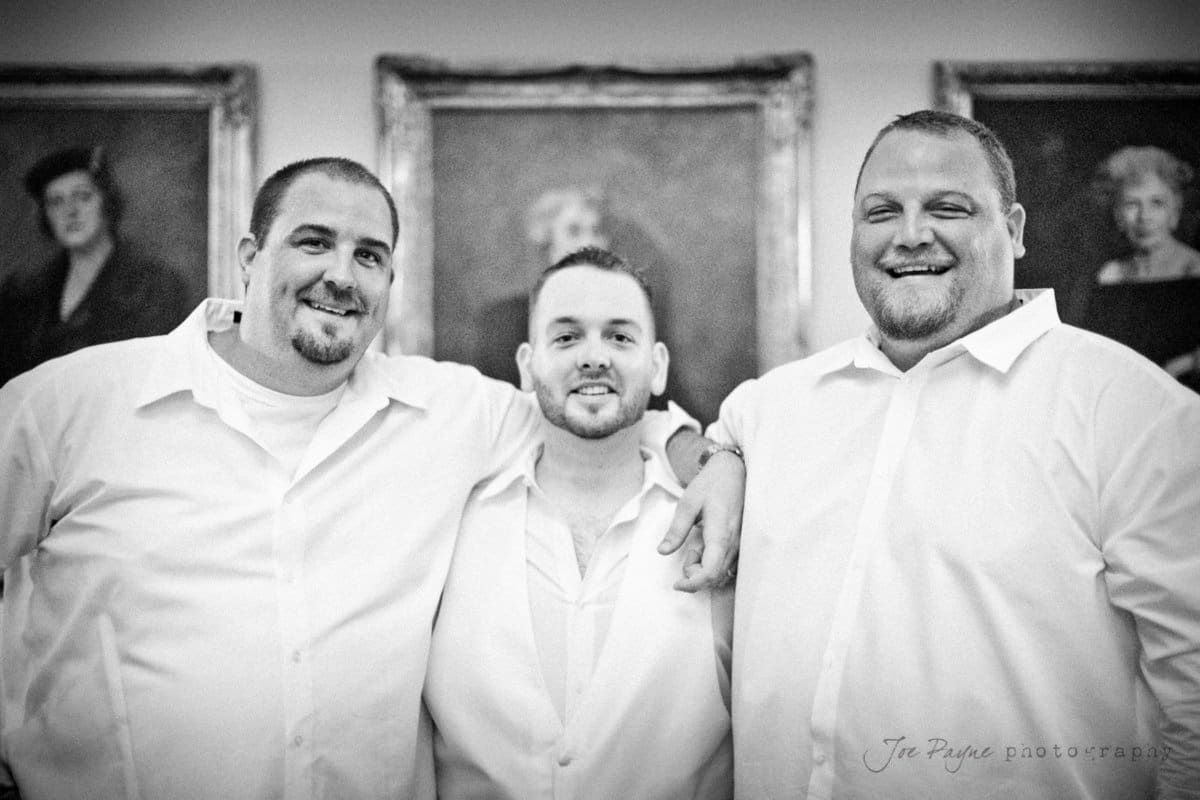 Three men in white shirts stand close together, smiling warmly at the camera. They are indoors, with framed portraits in the background, creating a cozy and friendly atmosphere.