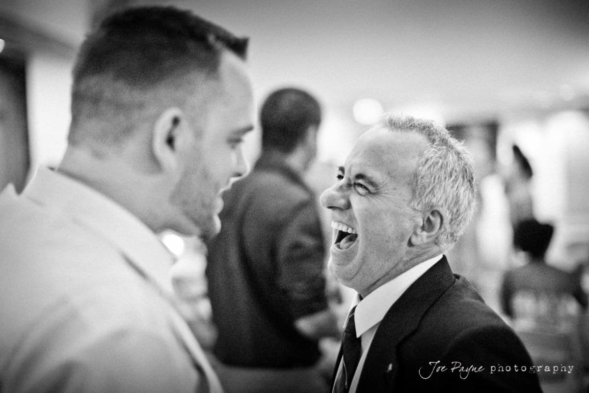 Two men facing each other, both smiling broadly. The man on the right is wearing a suit and tie, appearing to laugh joyfully. The background is blurred, indicating a lively atmosphere. Black and white photograph.