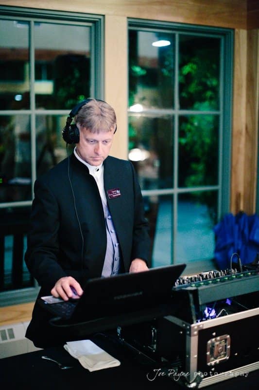 A DJ wearing headphones and a formal outfit is standing at a mixing console, focused on adjusting the controls. The setup includes a laptop and other audio equipment. He is in a room with large windows and soft lighting.