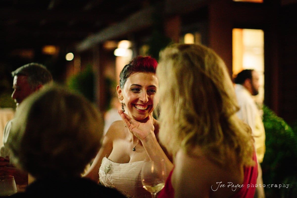 A festive gathering with friends. A woman with red hair, wearing a white dress, smiles brightly while engaging in conversation, surrounded by others holding drinks. The setting is warmly lit, creating an inviting atmosphere.