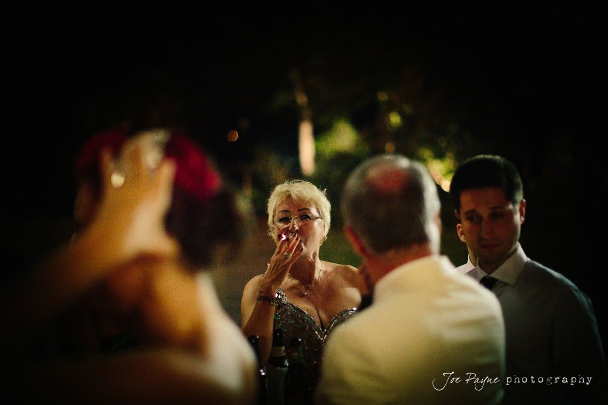 A group of people stands in semi-formal attire at a nighttime event. A woman in a sparkly dress is taking a drink, while others engage in conversation. Dim lighting and blurred background create an intimate atmosphere.