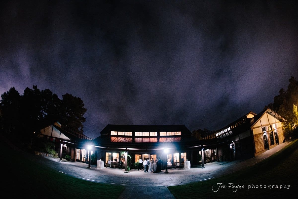 A nighttime photo of a large, well-lit building under a cloudy sky. The glow from the building illuminates the surrounding area, with trees visible in the background. Tables are set up outside, suggesting an event or gathering.