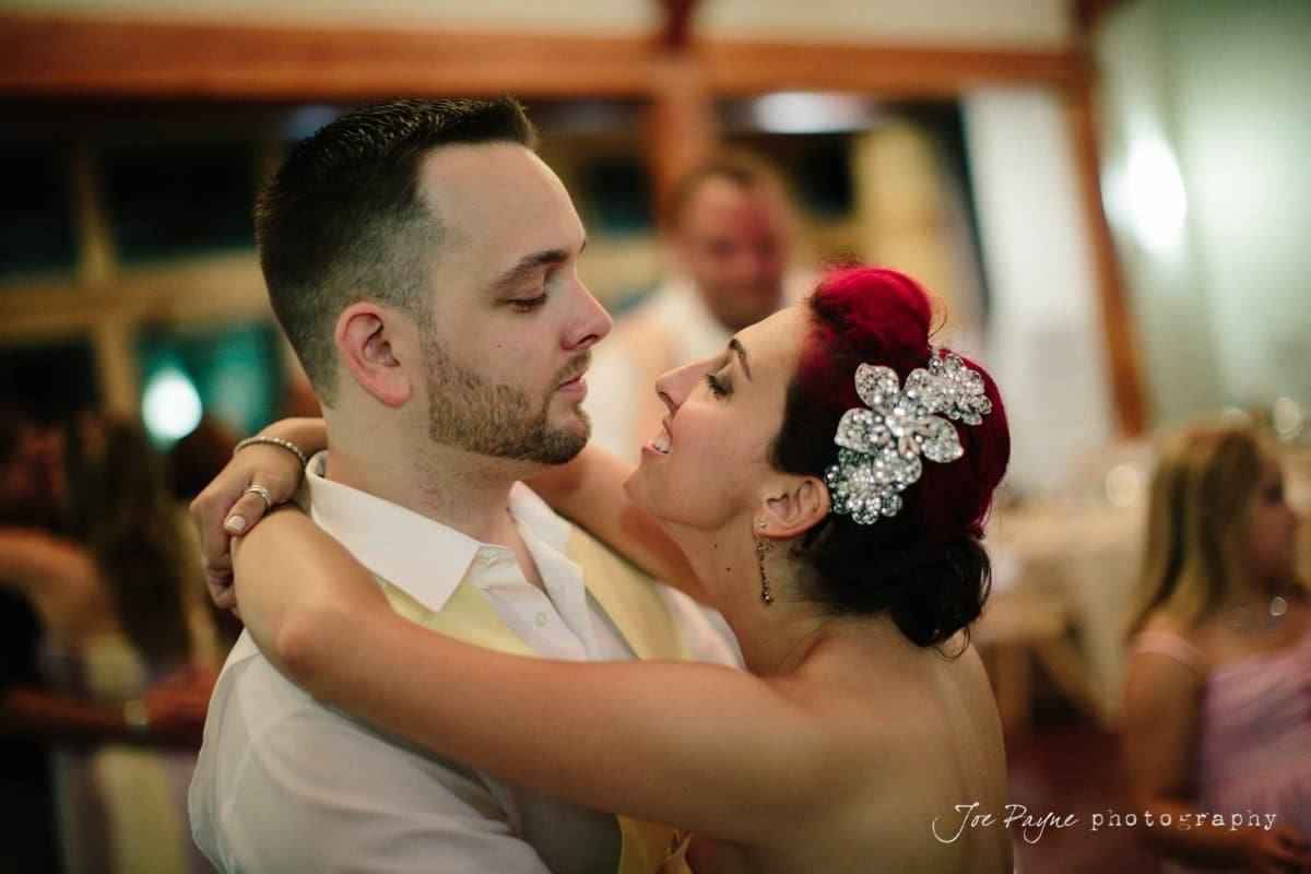 A couple is dancing closely at a formal event. The man is wearing a white shirt and a beige vest, while the woman, with a decorative hairpiece, embraces him, looking up with a joyful expression. The background is softly blurred with other guests visible.