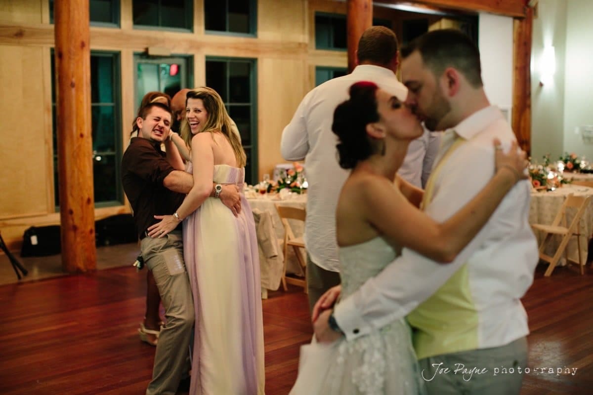 Two couples are dancing at a wedding reception. The couple in the foreground is kissing, while the couple behind them is smiling and having fun. The room is warmly lit with decorated tables in the background.