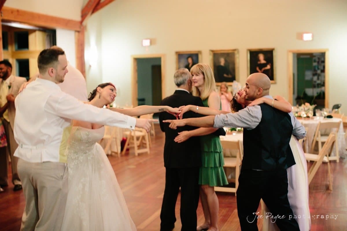 A joyful wedding scene with a bride and groom smiling, extending their arms toward each other on a dance floor. Guests dance around them, embracing and enjoying the festive atmosphere in a warmly lit venue.