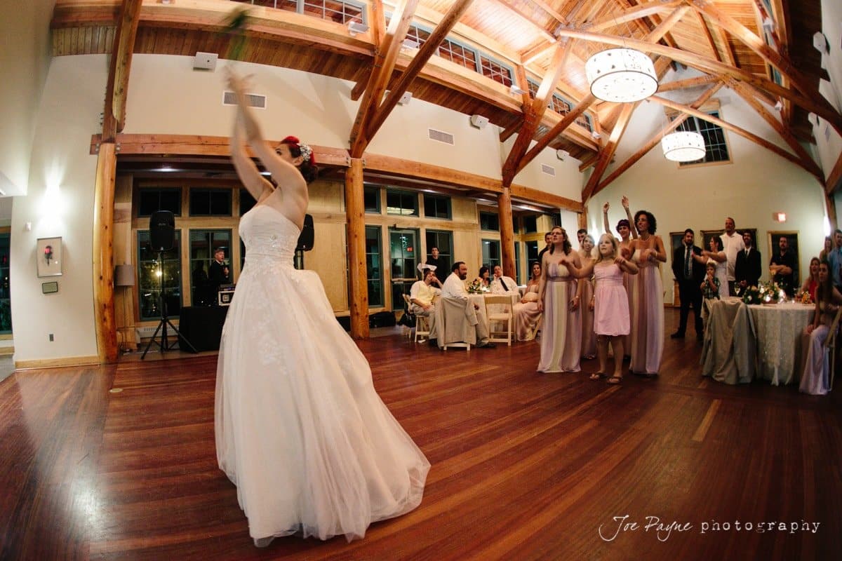 A bride in a white gown prepares to throw her bouquet to a group of eagerly awaiting bridesmaids in a spacious reception hall with wooden beams and chandeliers. Guests seated at tables watch the event unfold.