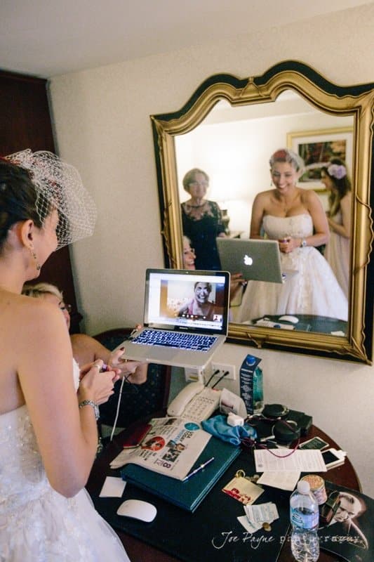 A bride in a wedding dress holds a phone while smiling at a laptop showing a video call. A mirror reflects her image and another woman in the background. The table is cluttered with various items.
