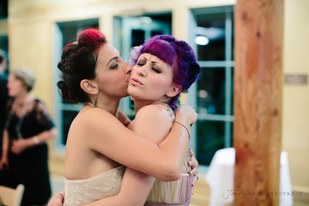 Two women with vibrant hairstyles embrace playfully indoors. One kisses the others cheek while the other makes a playful face. A wooden pillar and large windows are in the background.