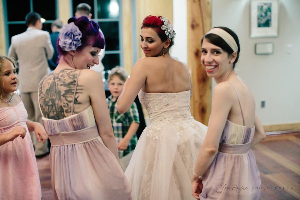 Three women in purple and white dresses, two with colorful hair, dance and smile at a wedding reception with children nearby. In the background, people are mingling and a little boy in a plaid shirt looks on.