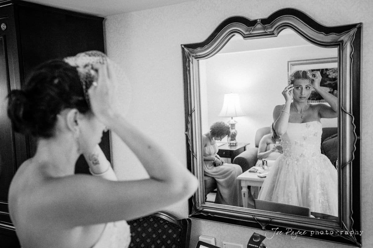 A bride adjusts her veil in front of a mirror. She is wearing a strapless wedding dress with lace details. In the background, people can be seen sitting on a couch. The reflection in the mirror shows her focused expression.