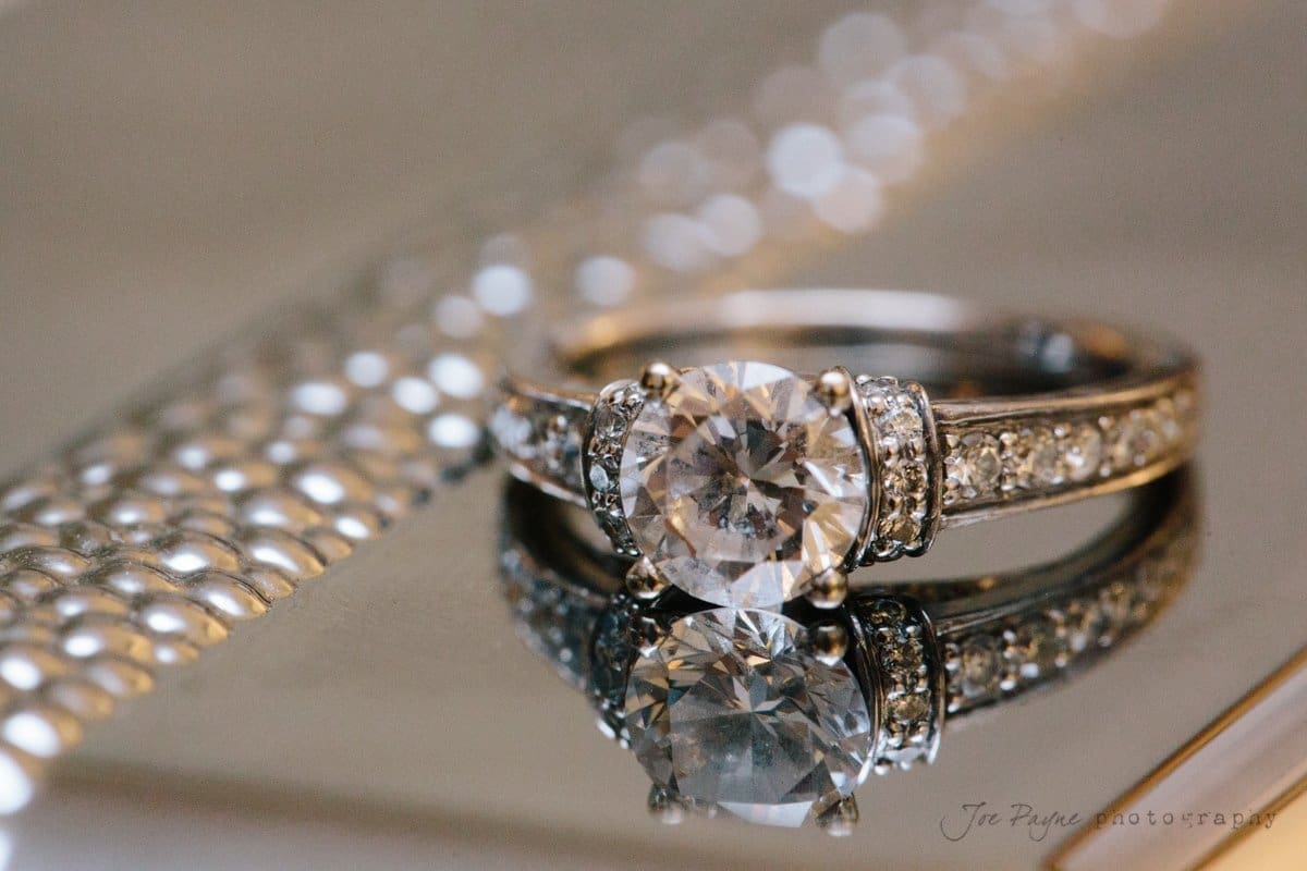 A close-up of an elegant diamond engagement ring with a large central stone, placed on a reflective surface. The band is adorned with smaller diamonds, and the image has a bokeh effect in the background.