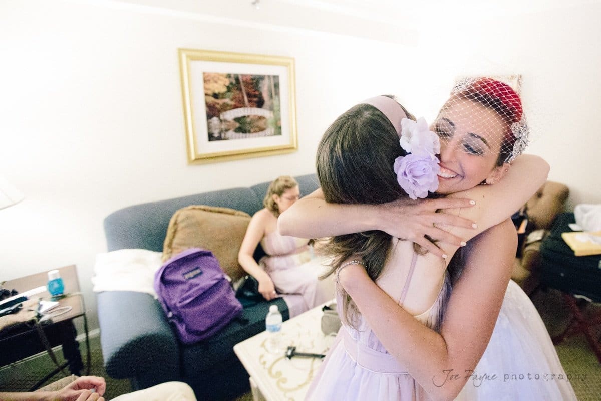 A bride with red hair and a birdcage veil hugs a bridesmaid in a room. The bridesmaid wears a lavender dress with a flower in her hair. Other people sit on a couch in the background. A framed landscape picture hangs on the wall.