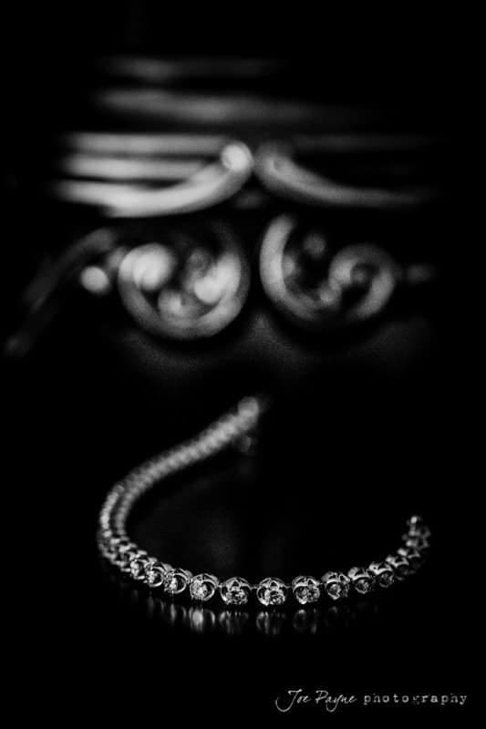 A curved bracelet with sparkling stones is placed on a reflective black surface. The background is blurred, featuring ornate, swirling designs in shades of gray. The image is in black and white.