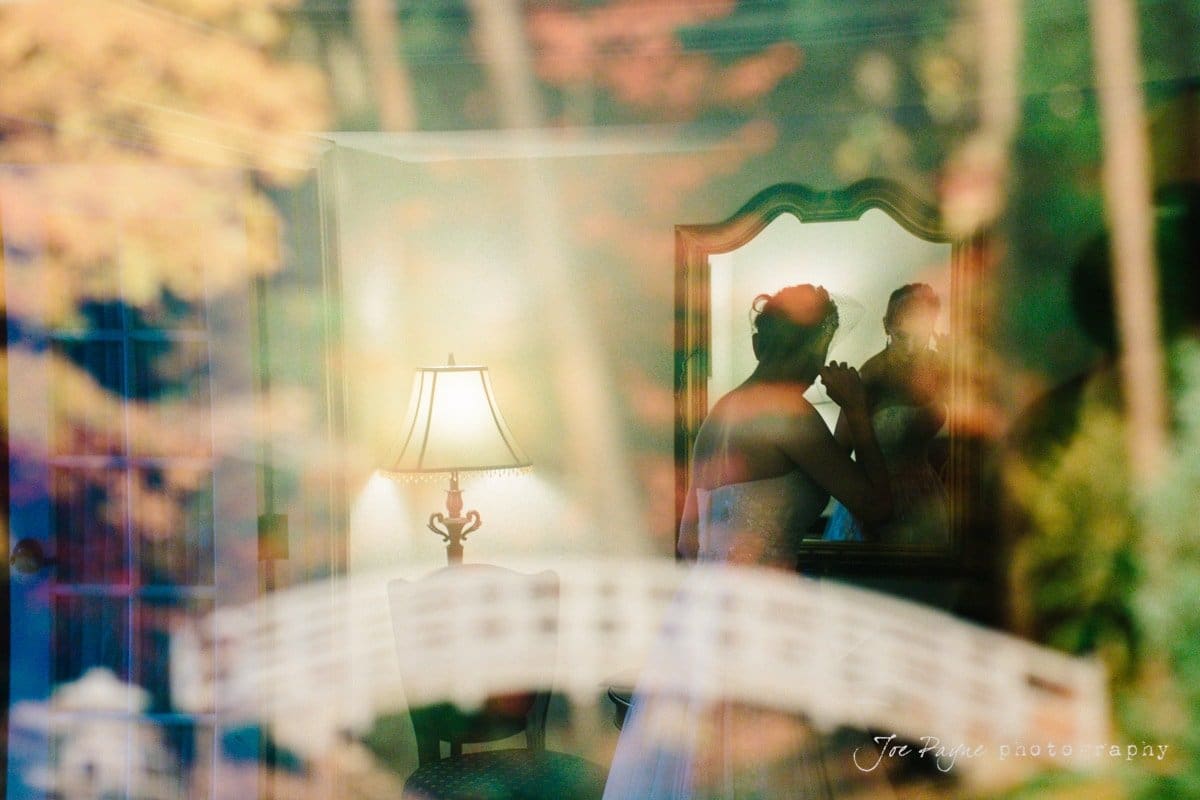 A bride is applying makeup in front of a mirror, with a vintage lamp nearby. The scene is artistically layered with reflections of autumn foliage, creating a dreamy and colorful effect.
